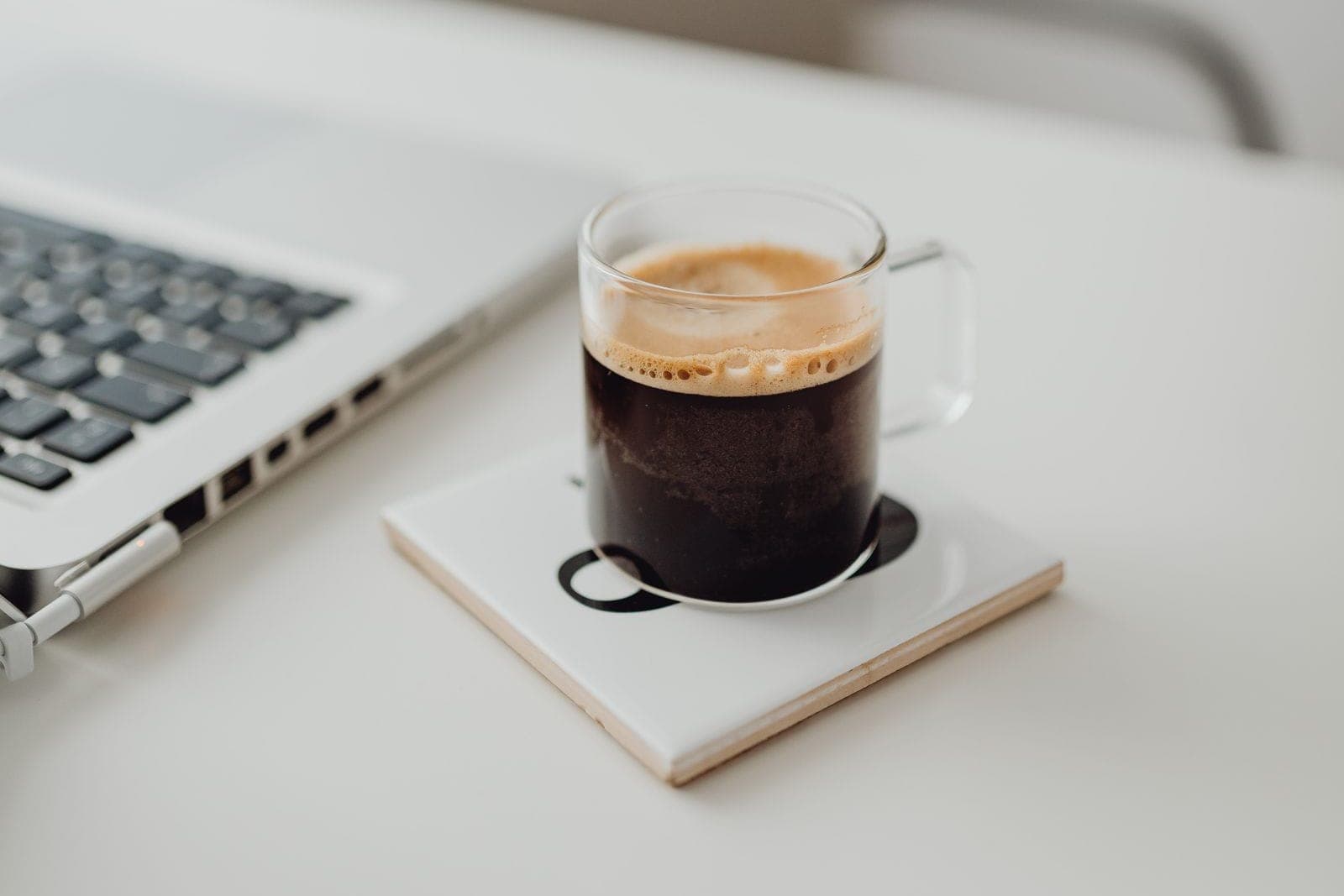 Glass cup of black coffee on a white desk beside a laptop