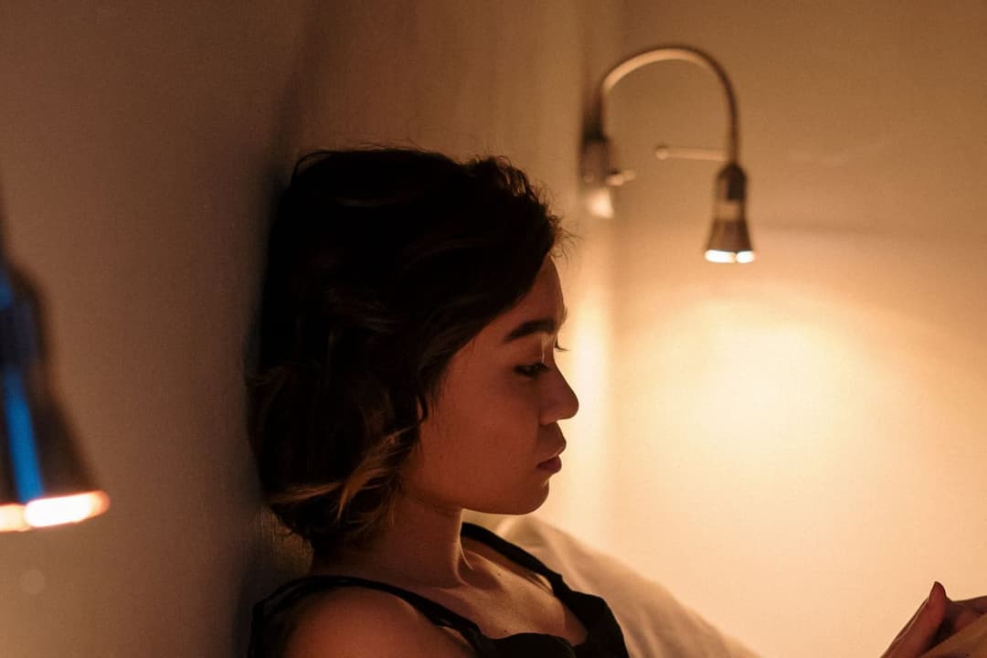 Woman sitting up in bed under a warm wall lamp